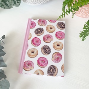 May include: A pink and white notebook with a donut pattern. The donuts are pink, brown, and white with sprinkles.