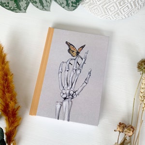 May include: A grey notebook with a skeleton hand and a butterfly on the cover. The notebook has a light brown spine.