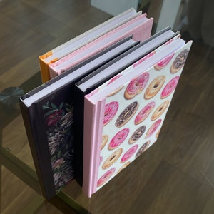 May include: Four different notebooks with colourful covers. The top notebook has a pink and white cover. The second notebook has a pink and white cover with a floral design. The third notebook has a black cover with a floral design. The bottom notebook has a white cover with a pink and brown donut pattern.