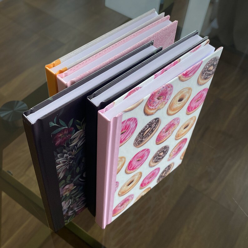 May include: Four different notebooks with colourful covers. The top notebook has a pink and white cover. The second notebook has a pink and white cover with a floral design. The third notebook has a black cover with a floral design. The bottom notebook has a white cover with a pink and brown donut pattern.