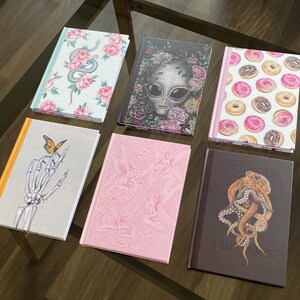 May include: Set of six hardcover journals with various illustrated covers. The covers feature a floral design with a snake, an alien, doughnuts, fairies, a skeleton hand holding a butterfly, and an octopus.