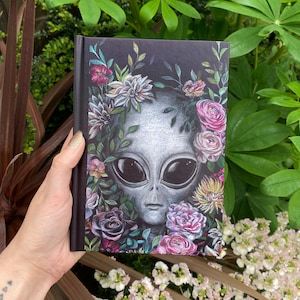 May include: A black journal with a gray alien face surrounded by colorful flowers. The alien has large eyes and a neutral expression. The flowers are a mix of pinks, purples, and greens.