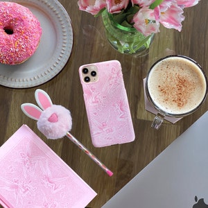May include: A pink iPhone case featuring a design of fairies and flowers. The case is on a wooden table with a doughnut, a cup of coffee, and a laptop.