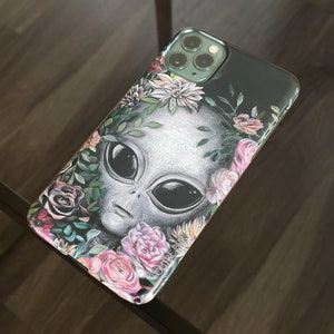 Op de afbeelding: Een zwarte telefoonhoes met een bloemenontwerp en een grijs alien gezicht. Het alien heeft grote ogen en een kleine mond. De bloemen zijn roze, wit en groen. De hoes heeft de tekst "OUIREFAN" onderaan gedrukt.