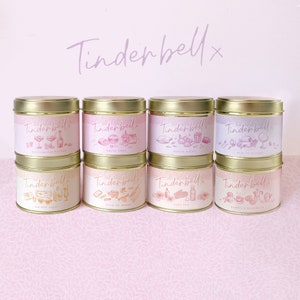 May include: Eight gold candle tins with pink and white labels. The labels feature illustrations of various activities and the text "Tinderbell" with a small "x" after it. The candle scents are: "Bottomless Brunch", "Drive Time", "Movie Night", "Pretty Rooms", "On The Hay Vacay", "Walk Up Shame", "Dark Tan", and "Coffee & Croissants".