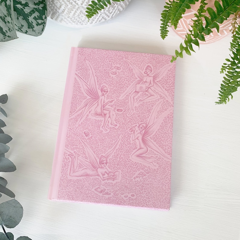 May include: A pink notebook featuring a repeating pattern of fairies with wings and sunglasses. The fairies are wearing various outfits and accessories, including a handbag, a book, and a cup.