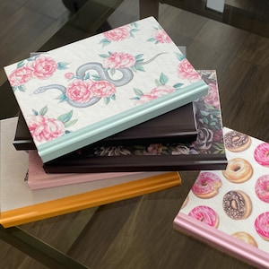 May include: A stack of five journals with different floral and patterned covers. The top journal has a light blue cover with a floral design and a snake. The second journal has a dark brown cover with a floral design. The third journal has a light pink cover with a floral design. The fourth journal has a light orange cover. The fifth journal has a white cover with a donut pattern.