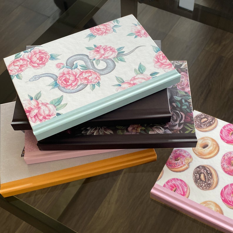 May include: A stack of five journals with different floral and patterned covers. The top journal has a light blue cover with a floral design and a snake. The second journal has a dark brown cover with a floral design. The third journal has a light pink cover with a floral design. The fourth journal has a light orange cover. The fifth journal has a white cover with a donut pattern.