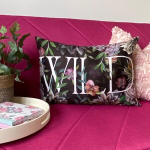 May include: A black pillow with a floral pattern and the word "WILD" printed in white. The pillow is on a pink couch.