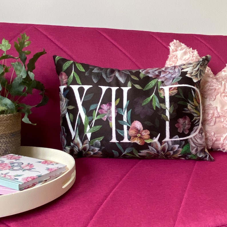 May include: A black pillow with a floral pattern and the word "WILD" printed in white. The pillow is on a pink couch.