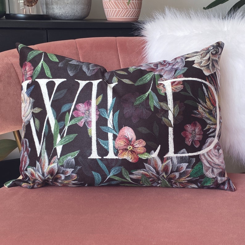 May include: A black pillow with a floral print and the word "WILD" in white lettering. The pillow is on a pink velvet couch.