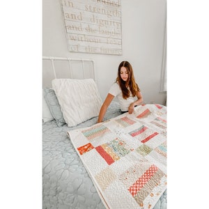 May include: A colourful patchwork quilt featuring squares of patterned fabric in red, pink, yellow, and green, placed on a bed with a light blue floral bedspread. The quilt has white quilted details. A sign above the bed reads "She is clothed in strength and dignity and she laughs without fear of the future."