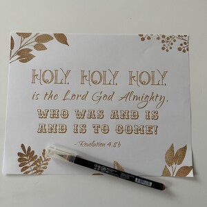 May include: White paper print with gold text that reads "HOLY, HOLY, HOLY, is the Lord God Almighty, WHO WAS AND IS AND IS TO COME!" with "Revelation 4:8b" below. Gold leaf designs are in the corners. A brown eyeliner pencil with a clear cap rests on the paper.