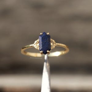 Peut inclure: Une bague en or ornée d'une pierre précieuse rectangulaire bleu foncé. La monture de la bague présente un motif délicat en forme de feuille de chaque côté de la pierre. La bague est tenue par une pince, mettant en valeur ses détails.