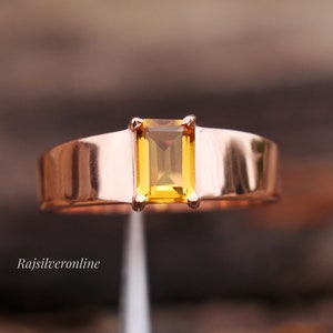 Op de afbeelding: Een gouden ring met een rechthoekige, gele edelsteen in het midden. De ring ligt op een witte achtergrond.