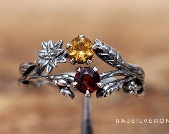 Puede incluir: Un anillo de plata con un delicado diseño floral que presenta tres piedras preciosas: un granate rojo, un citrino amarillo y una piedra blanca. El anillo está colocado sobre una superficie de madera.