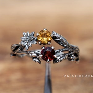 Könnte beinhalten: Ein silberner Ring mit einem zarten floralen Design, das mit drei Edelsteinen verziert ist: einem roten Granat, einem gelben Citrin und einem weißen Stein. Der Ring liegt auf einer Holzoberfläche.