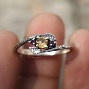 Op de afbeelding: Een zilveren ring met een op de natuur geïnspireerd ontwerp. De ring heeft een centrale gele edelsteen, geflankeerd door twee kleinere paarse edelstenen. De band heeft een getextureerd, takachtig uiterlijk, met blad details.