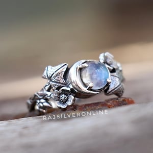 Può includere: Un anello d'argento con una grande gemma rotonda e iridescente. L'anello presenta intricati disegni floreali e di foglie sulla fascia. Il testo "RAJSILVERONLINE" è visibile.