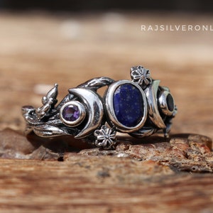 May include: A silver ring with a crescent moon design, featuring a blue lapis lazuli gemstone, a purple amethyst gemstone, and a black onyx gemstone. The ring is adorned with delicate floral details.