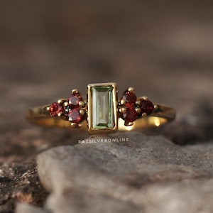 May include: A gold ring with a central rectangular, pale green gemstone, complemented by clusters of small, red gemstones. The ring's slender band is set against a blurred, natural backdrop.