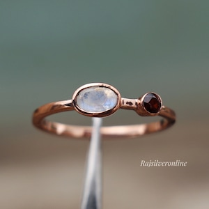 May include: A delicate rose gold ring featuring a white moonstone and a small red garnet. The ring is displayed against a silver background.