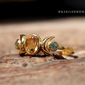 Könnte beinhalten: Goldring mit drei Mondsicheln und einem großen ovalen Topas-Edelstein. Der Ring liegt auf einer Holzoberfläche.