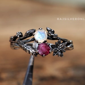 May include: A silver ring with a floral design, featuring a round, light blue gemstone and a round, red gemstone. The ring has a detailed, leaf-like pattern and a small flower on the side. The text "rajsilveronline" is visible.