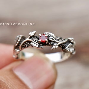 May include: A silver ring with a red gemstone set in a vine design. The ring is made of sterling silver and features a delicate vine design with leaves and a single red gemstone.