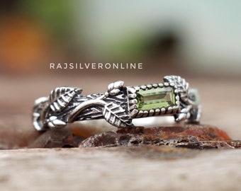 Peut inclure: Une bague en argent avec une pierre précieuse verte sertie dans un motif de feuilles. La bague est posée sur une surface en bois.