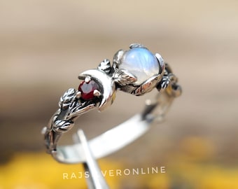 May include: A silver ring featuring a moon and leaf design, with a round, iridescent moonstone and a small, red gemstone. The ring has a detailed, nature-inspired aesthetic. The text "RAJSVERONLINE" is visible.