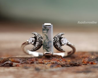 May include: A silver ring with a green and white agate stone and two crescent moon accents set with small clear stones. The ring is on a wooden surface.