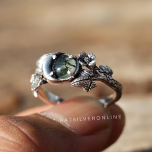 Könnte beinhalten: Ein silberner Ring mit einem grünen Moosachat-Stein, der in einem Halbmond-Design gefasst ist. Der Ring ist mit zarten floralen und Blatt-Akzenten verziert.