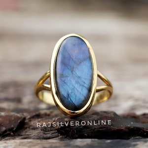 May include: A gold-toned ring featuring an oval-shaped, blue-hued gemstone. The gemstone is set in a gold bezel, and the band has a split design. The text "RAJSILVERONLINE" is visible at the bottom.