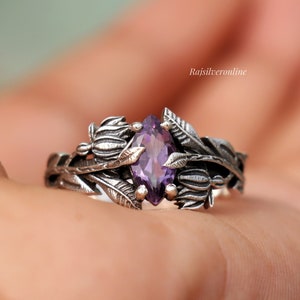 May include: A silver ring with a purple gemstone set in a floral design. The ring is on a hand.