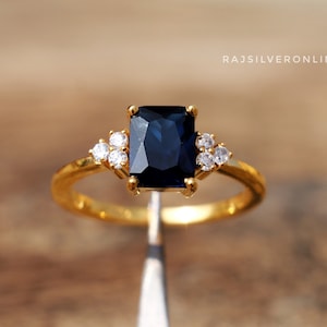 May include: A gold ring with a large, dark blue, rectangular-shaped gemstone surrounded by smaller, clear gemstones.