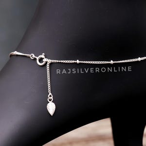 Puede incluir: Una delicada tobillera de plata con un dije de hoja. La tobillera presenta una fina cadena con pequeños detalles de cuentas y un colgante de hoja. El cierre es visible y el texto "RAJSILVERONLINE" está impreso.