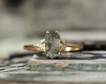 May include: A gold ring featuring a hexagonal moss agate gemstone with green inclusions. The band is designed with delicate leaf-shaped metal details on both sides of the stone, which is held in place by a six-prong setting in a polished gold finish.