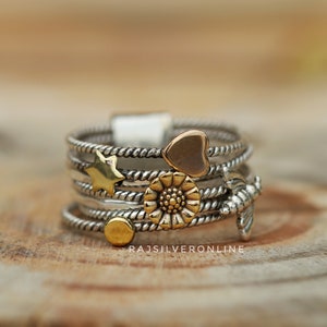 May include: A silver ring with a twisted band and multiple charms. The charms include a heart, a star, a flower, and a bird. The ring is on a wooden surface.