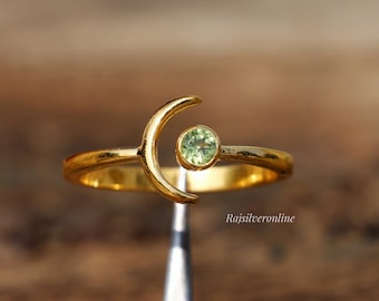 May include: Gold ring with a crescent moon design and a small green gemstone. The ring is on a display stand.