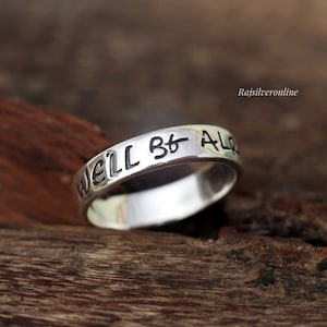 Op de afbeelding: Een zilveren ring met de gegraveerde tekst "WELL BE AL" in zwarte letters. De ring is geplaatst tegen een rustieke houten achtergrond, die het gepolijste oppervlak en het contrast van de donkere tekst benadrukt.