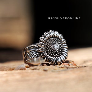 Puede incluir: Un anillo de plata con un diseño de girasol. El girasol tiene un centro texturizado y está rodeado de pétalos. La banda del anillo también está texturizada y tiene un diseño floral.