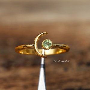 May include: Gold ring with a crescent moon design and a small green gemstone. The ring is on a display stand.