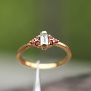 May include: A gold ring featuring a rectangular, light-colored center stone flanked by smaller, round red stones. The ring has a delicate band and is set against a blurred green background. The ring is held by a pair of tweezers.