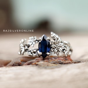 May include: A silver ring featuring a blue gemstone surrounded by floral details. The ring has a marquise-shaped gemstone and delicate floral accents.