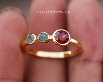 May include: Gold ring with three gemstones. The center stone is red, and the two smaller stones on either side are green.