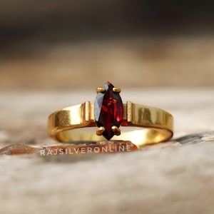 Peut inclure: Une bague en or avec une pierre précieuse rouge en forme de marquise. La bague est posée sur une surface en bois.