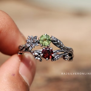 Peut inclure: Une bague en argent avec un délicat motif floral orné d'un grenat rouge, d'une péridot verte et d'une fleur blanche.