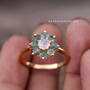 Puede incluir: Un anillo de oro con una gran piedra de luna redonda y blanca rodeada de pequeñas piedras preciosas redondas verdes. El anillo está engastado en un diseño con forma de flor.