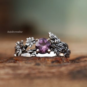 Puede incluir: Un anillo de plata con una piedra preciosa púrpura rodeada de delicados diseños florales. El anillo está colocado sobre una superficie de madera rústica.